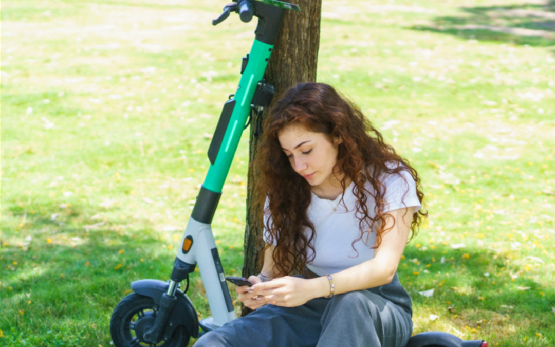 Photo of a girl sat under a tree next to a turquoise electric scooter.